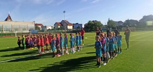 Na boisku piłkarskim stoją drużyny czterech zespołów wraz ze swoimi trenerami