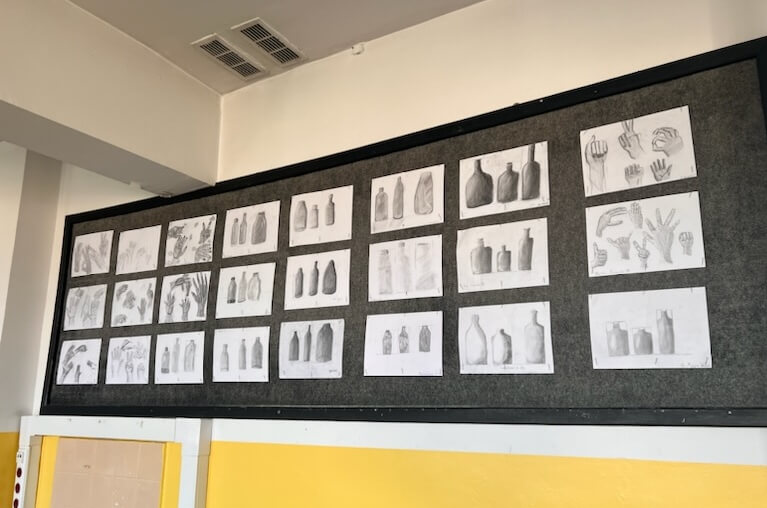 Na tablicy wiszą rysunki uczniów przedstawiające martwą naturę (butelki) oraz dłonie w różnej pozycji.