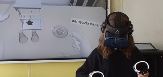 uczennica stoi przed tablicą mając założone na oczy gogle vr , trzyma w rękach joystick by wykonać określone zadanie.