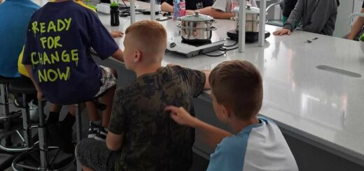 Uczniowie siedzą przy białych stołach , na których stoją garnki na kuchenkach . Słuchają Pani prowadzącej zajęcia.