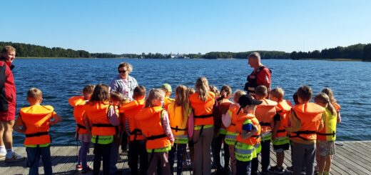 Dzieci ubrane w kamizelki ratunkowe stoją na pomoście i słuchają ratownika WOPR.