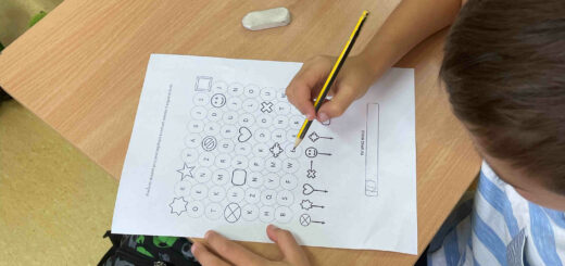 Uczeń siedzący przy stoliku próbuje odkodować informację. Trzyma przed sobą kartkę z kodami , w ręku ma ołówek.