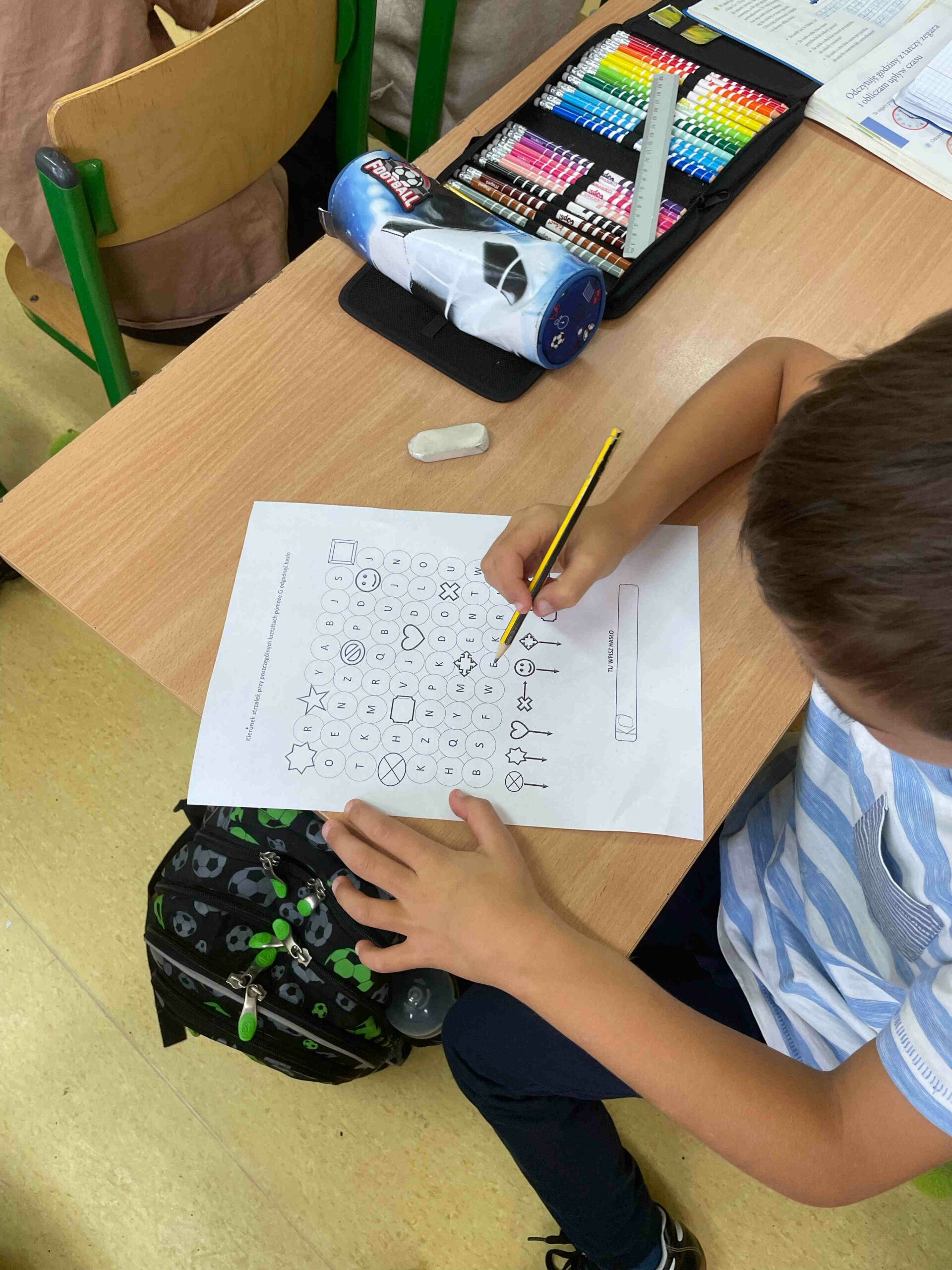 Uczeń siedzący przy stoliku próbuje odkodować informację. Trzyma przed sobą kartkę z kodami , w ręku ma ołówek.