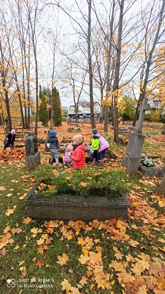 Grupka dzieci porządkuje zapomniany grób na cmentarzu na dajtkach w Olsztynie. Na grobach leży mnóstwo liści w wielu odcieniach czerwieni.