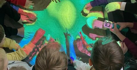 Dzieci siedząc na podłodze wysunęły ręce pod żółto - zielono - niebieskie światło.