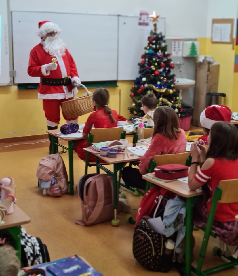 Przed uczniami siedzącymi w ławkach stoi Mikołaj z wielkim koszem wiklinowym. W rogu stoi duża choinka przystrojona w bombki.