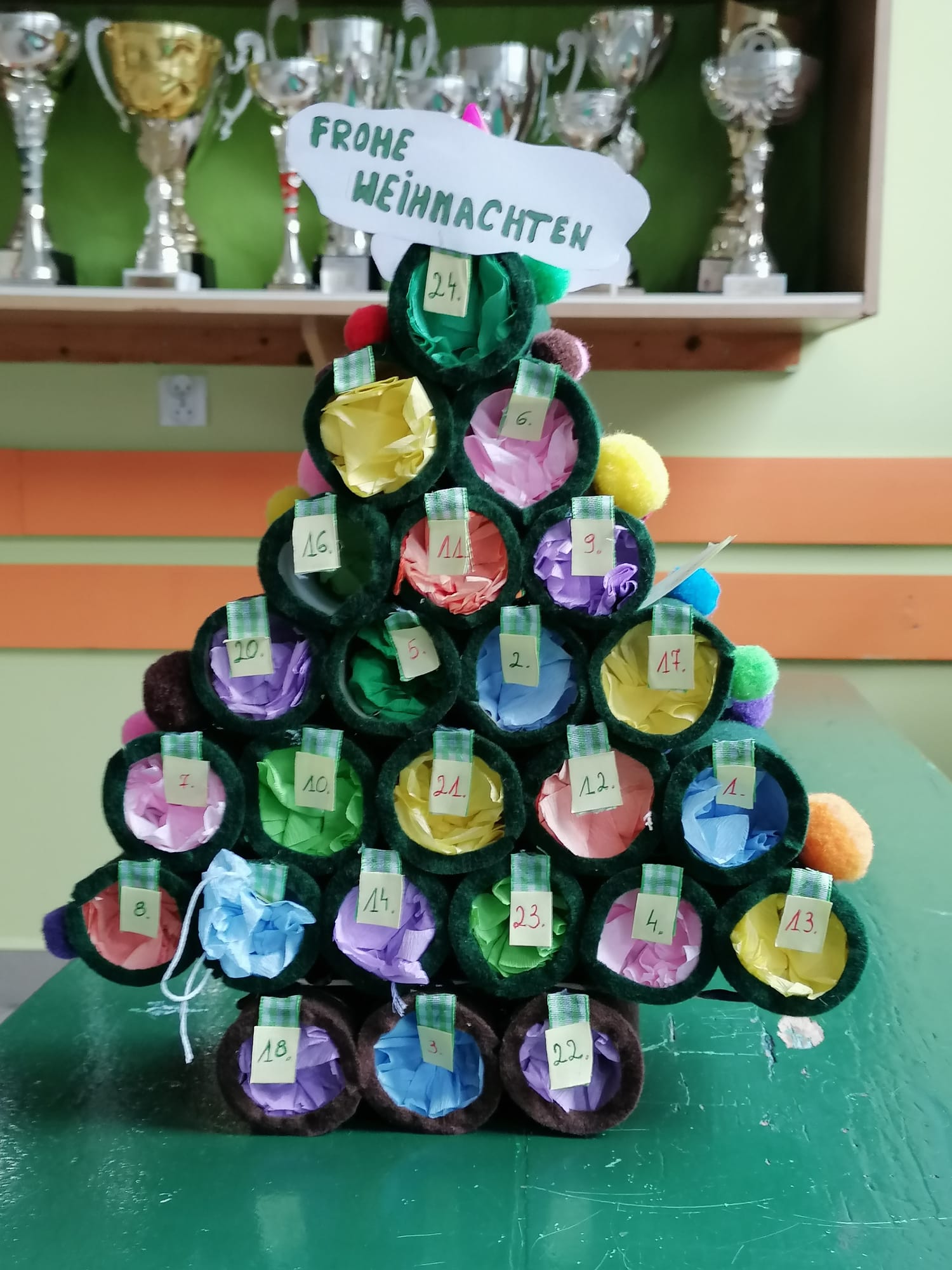 Kalendarz adwentowy w kształcie choinki, Okrągłe rulony pomalowane na zielono, otwór zakryty kolorowymi bibułkami i numerami. Na czubku choinki napis - Frohe Weihnachten.