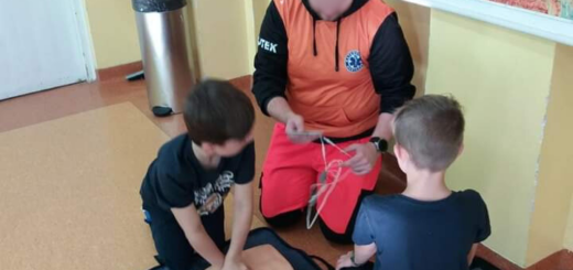 Na podłodze w klasie klęczy ratownik oraz dwóch uczniów, którzy uczą się udzielać pierwszą pomoc na fantomie.