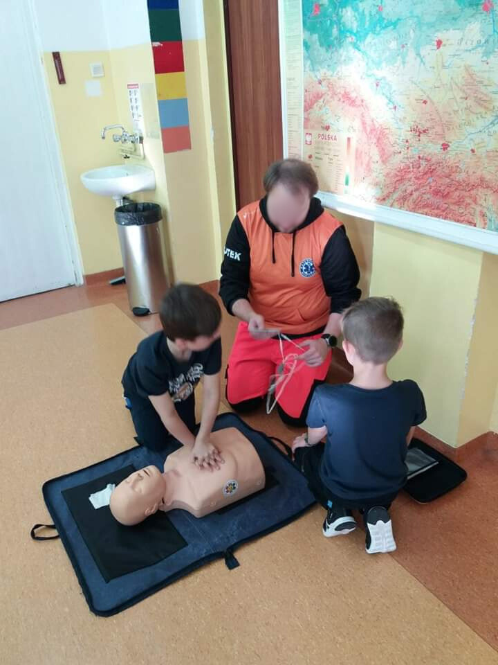 Na podłodze w klasie klęczy ratownik oraz dwóch uczniów, którzy uczą się udzielać pierwszą pomoc na fantomie.