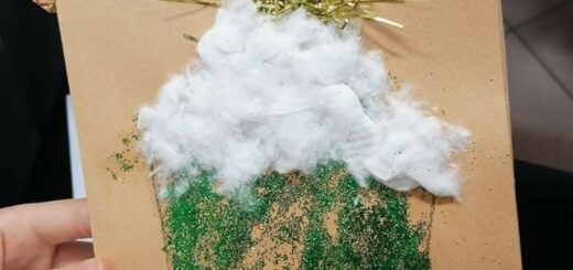 osoba trzyma beżową kartkę świąteczną na której znajduje się zielony domek z białym dachem, nad którym świeci złota gwiazda.