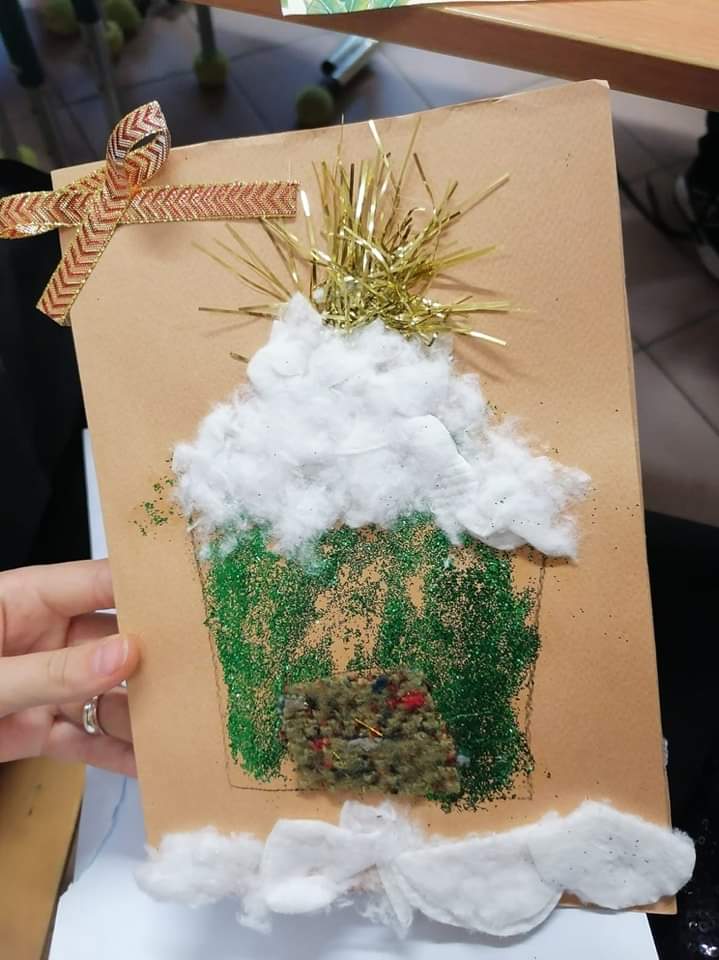 osoba trzyma beżową kartkę świąteczną na której znajduje się zielony domek z białym dachem, nad którym świeci złota gwiazda.