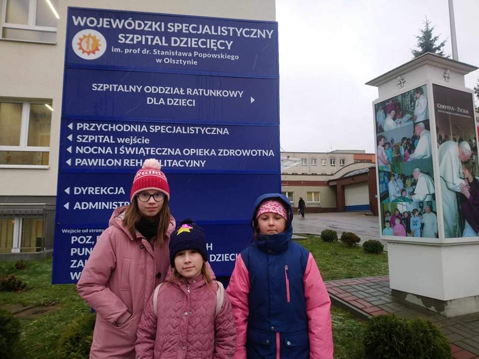 Trzy ubrane w ciepłe kurtki i czapki dziewczyny stoją na tle niebieskiej tablicy informacyjnej szpitala w Olsztynie.