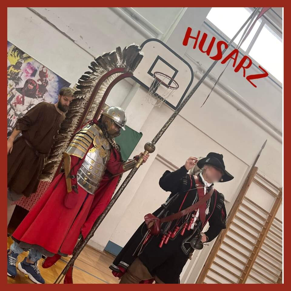 Na zdjęciu stoją dwaj mężczyźni jeden ubrany w zbroję siedemnastowiecznego rycerza z husarii drugi ubrany w strój szwedzkiego żołnierza - czarny kapelusz i czarny płaszcz. 