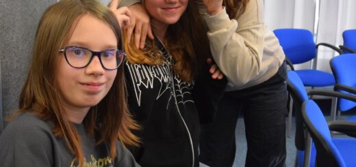 Na zdjęciu znajdują się trzy dziewczynki pierwsza w okularach siedzi, dwie pozostałe stoją przytulając się do siebie.