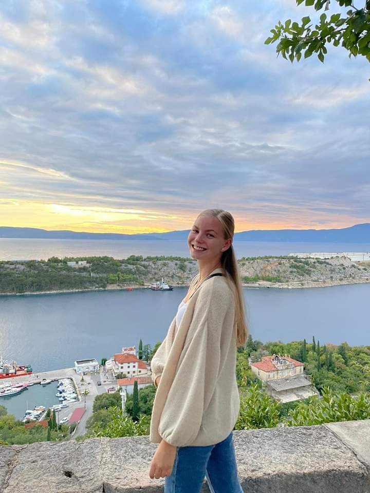 Na wzgórzu nad zatoką o zachodzie słońca stoi blondynka z długimi włosami. Na jej twarzy rysuje się uśmiech, ubrana jest w białą bluzę i jeansy.