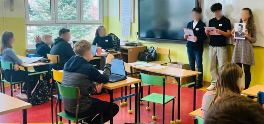 Troje uczniów stoi pod tablicą . Trzymają w rękach ilustracje. Dzieci siedzą w ławkach i słuchają przemawiających uczniów.