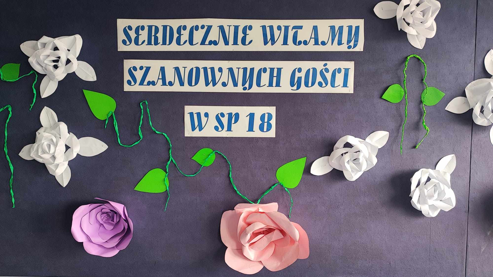 Na szarym tle niebieski napis  napis - Serdecznie witamy w szanownych gości w SP 18, wokół napisu znajdują się przyczepione kolorowe kwiaty.
