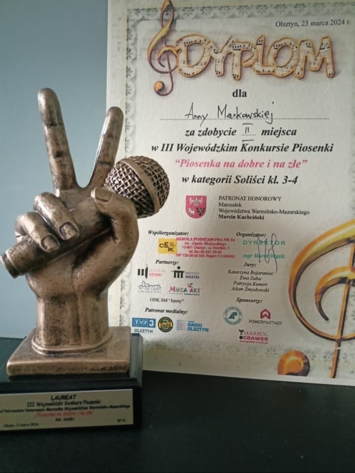 Na zdjęciu stoi statuetka jest to ręka trzymająca mikrofon . Za nią stoi dyplom .