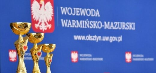 na niebieskim tle z białym napisem Wojewoda Warmińsko - Mazurski i godłem Polski stoją trzy złote puchary z czerwonym serduszkiem