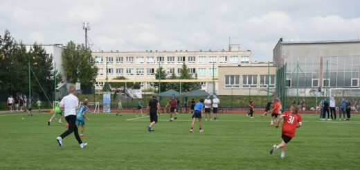 Na boisku grają uczniowie w piłkę nożną. W oddali widać obserwujących mecz. 