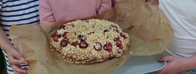 Dzieci trzymają na swoich rękach upieczone okrągłe  ciasto drożdżowe z owocami.