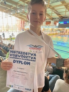 Na zdjęciu chłopiec w białej  koszulce w jednej ręce trzyma dyplom w drugiej zdobyty medal.