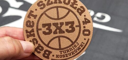 Na tle boiska sportowego dłoń trzyma drewniany medal z napisem Basket Szkoła 4.0; 3x3; Turniej koszykówki.