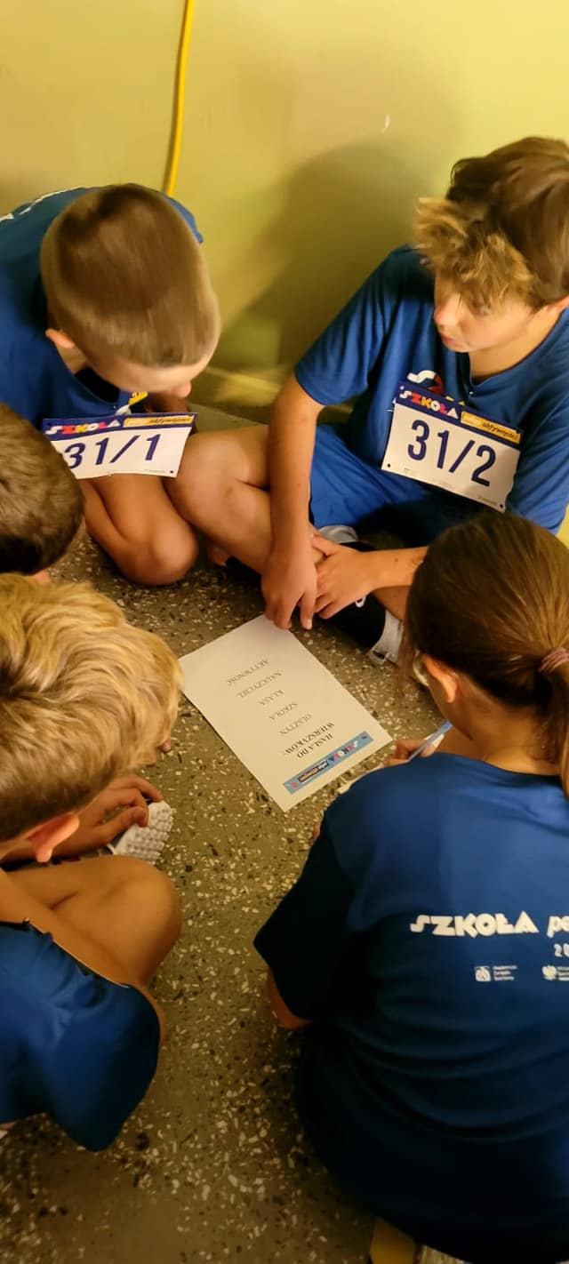 Zawodnicy w niebieskich koszulkach z numerami startowymi siedzą na podłodze i dyskutują jak rozwiązać dane zadanie.