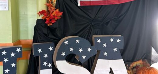 Na granatowym tle stoją duże litery USA w kolorach granatu , bieli i czerwieni. obok leżą pomarańczowe dynie . Nad nimi wisi flaga USA