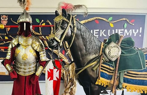 Na zdjęciu widoczna jest zbroja husarza srebrno - złota i bordowym ubraniem a także hełm. Obok zbroi stoi czarna figura konia również osiodłanego wg wzorców z epoki.