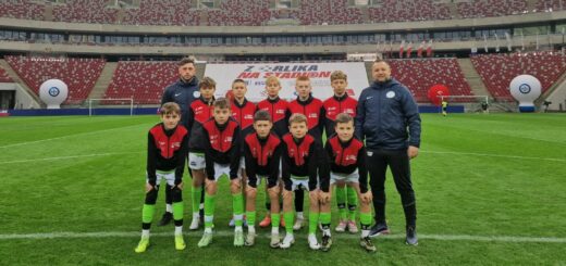 W tle zdjęcia trybuny stadionu Narodowego w Warszawie. Na pierwszym planie stoi 10 chłopców, młodych piłkarzy , ubranych w granatowo -czerwone bluzy. Z obu stron stoją dwaj trenerzy