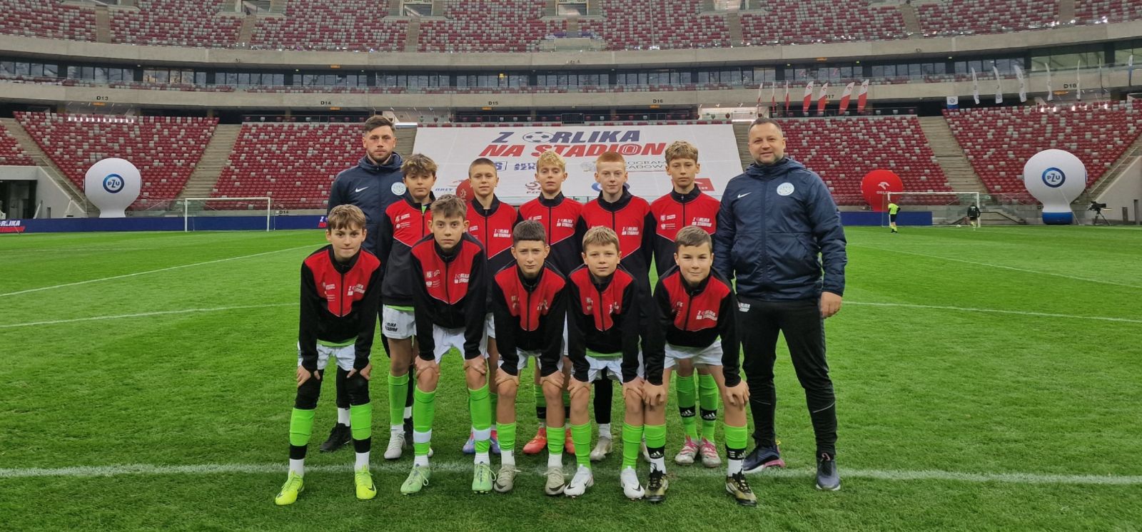 W tle zdjęcia trybuny stadionu Narodowego w Warszawie. Na pierwszym planie stoi 10 chłopców, młodych piłkarzy , ubranych w granatowo -czerwone bluzy. Z obu stron stoją dwaj trenerzy