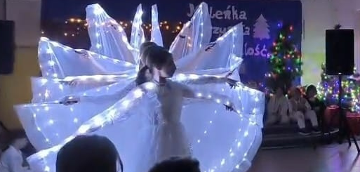 Dzieci tańczą ubrane w białe stroje , na rękach mają przyczepione  skrzydła ze światełkami ledowymi.